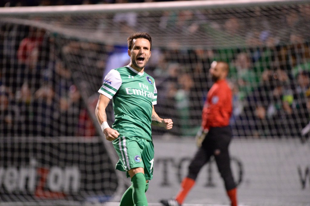 Soccer player in a green kit celebrating a goal with a clenched fist near the goal, with the goalkeeper and crowd visible in the background.